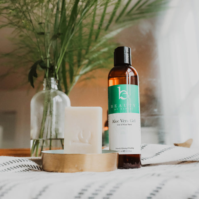 A Beauty by Earth Aloe Vera Gel bottle sits next to a soap bar in a wooden dish on striped fabric, embodying organic skincare. A clear vase with green foliage softly blurs in the background.