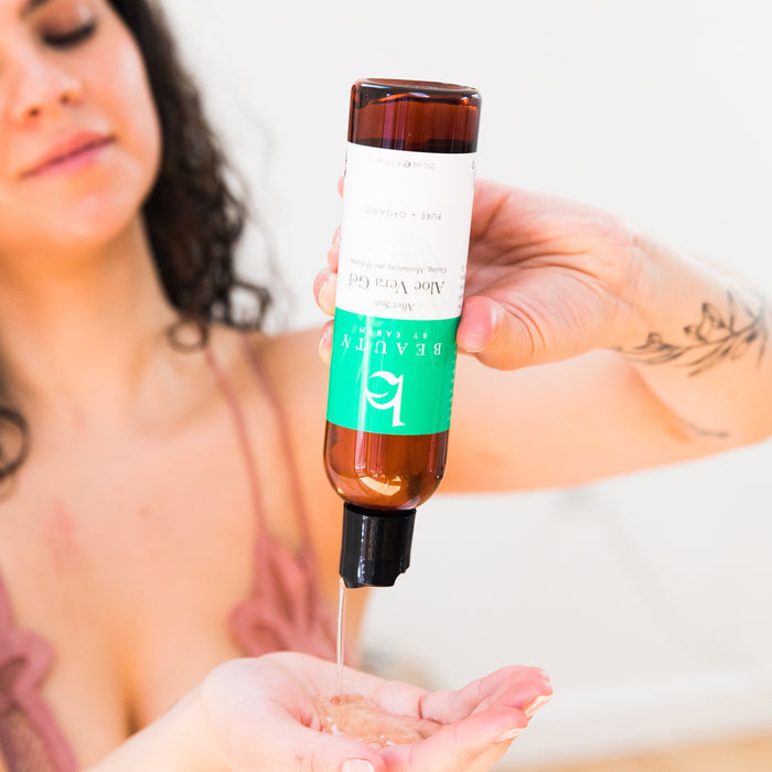 A person with long dark hair, wearing a light pink top, dispenses Beauty by Earths Aloe Vera Gel from a brown bottle with a green label and white text, indicating it as an organic skincare product.