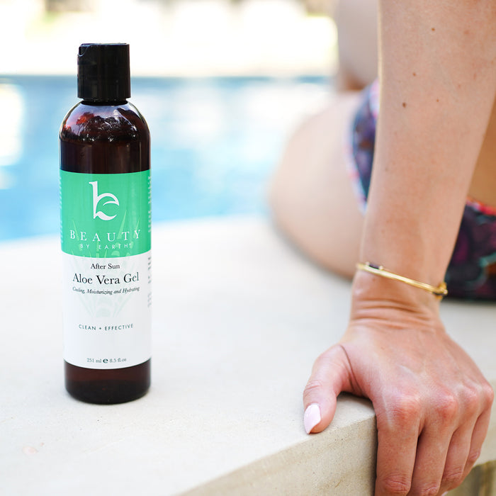 A bottle of Beauty by Earths Aloe Vera Gel rests on a poolside ledge. A person in vibrant swimwear casually extends a hand, capturing the essence of natural personal care. The shimmering pool water in the background completes this serene, summery tableau.