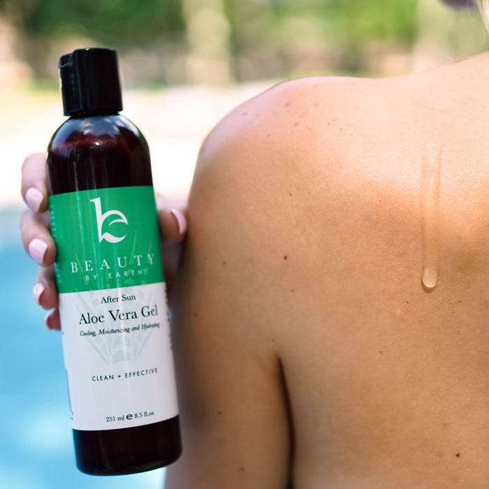 A person holds Beauty by Earth Aloe Vera Gel near their shoulder, revealing the After Sun Aloe Vera Gel and Clean + Effective labels. A glistening drop of this organic skincare solution is visible on their skin, with a blurry pool in the background.