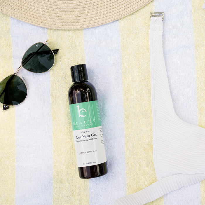 A beach scene featuring Beauty by Earths Aloe Vera Gel on a striped towel next to black sunglasses and a white sun hat, with part of a white swimsuit visible, suggesting the embrace of natural personal care on a sunny day at the beach.