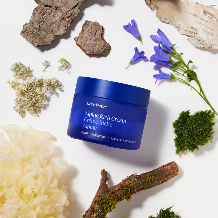 A blue jar of Ursa Major Alpine Rich Cream, a moisturizer for skin barrier support, sits on a white background surrounded by bark, moss, purple flowers, lichen, and white fungus.