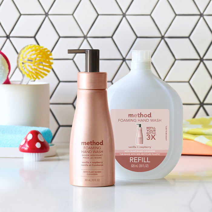 Aluminum Foaming Hand Soap by method displayed beside a refill pack on a bathroom counter, set against a white hexagonal tile wall with a mushroom-shaped decoration. Vanilla and raspberry scent makes it perfect for sensitive skin.