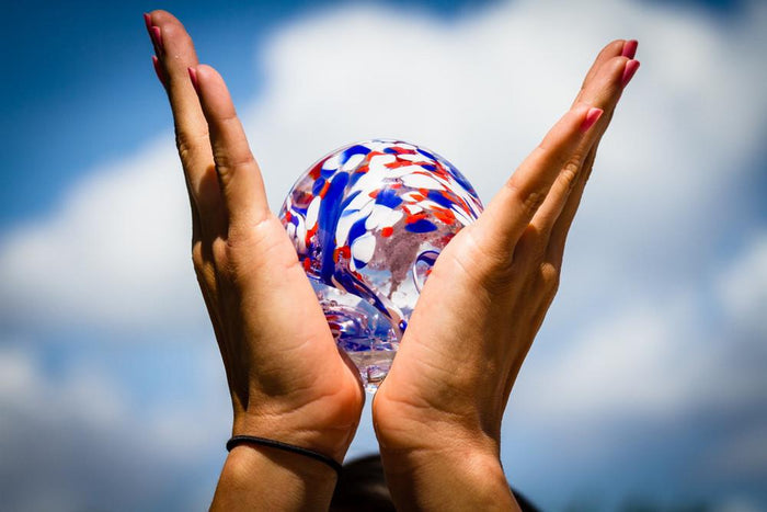 Hands cradle ash infused American Confetti Orb outdoors against a bright blue sky.