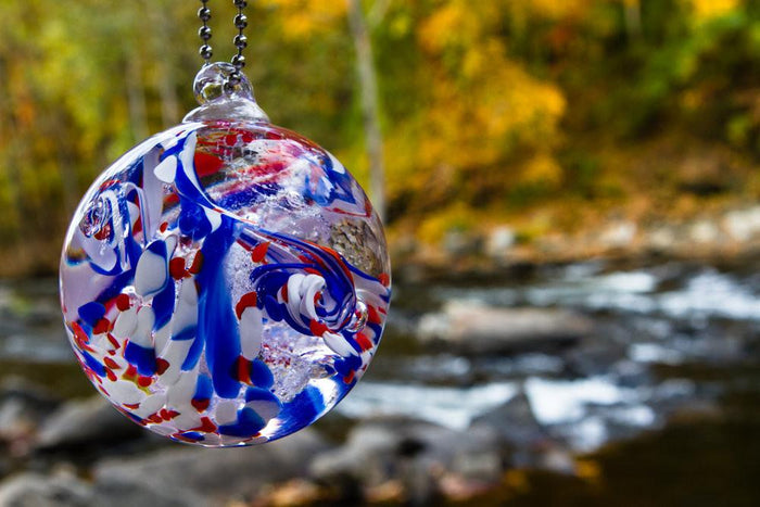 American Confetti Orb with a chain, vivid red white and blue swirls and ashes infused, outdoor blurred background.