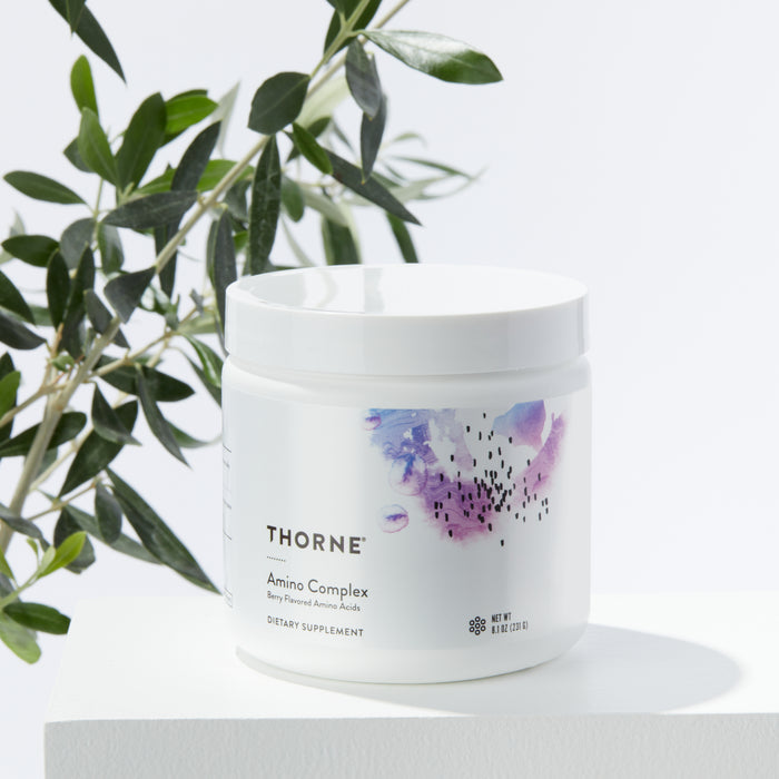 A white Thorne Amino Complex dietary supplement jar with essential amino acids rests on a pristine white surface. The label features a purple and pink abstract design, complemented by green leaves in the background for a natural touch.
