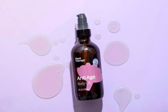 On a light purple surface lies a dark brown Anti Age Body Oil bottle by Plant Therapy, featuring a pump dispenser. Surrounding drops of essential oil blend promise youthful, glowing skin.