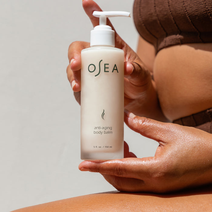 A person with glowing skin holds OSEA Anti-Aging Body Balm with a white pump, highlighting its formula that supports skin elasticity. The neutral background and brown textured top complete the scene.