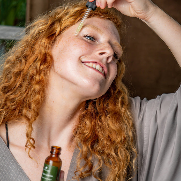 A person with long curly red hair wears a gray shirt and smiles as they happily apply Tree To Tubs Anti Aging Green Tea, Hyaluronic & Retinol Serum for Face using a dropper from a small brown bottle.
