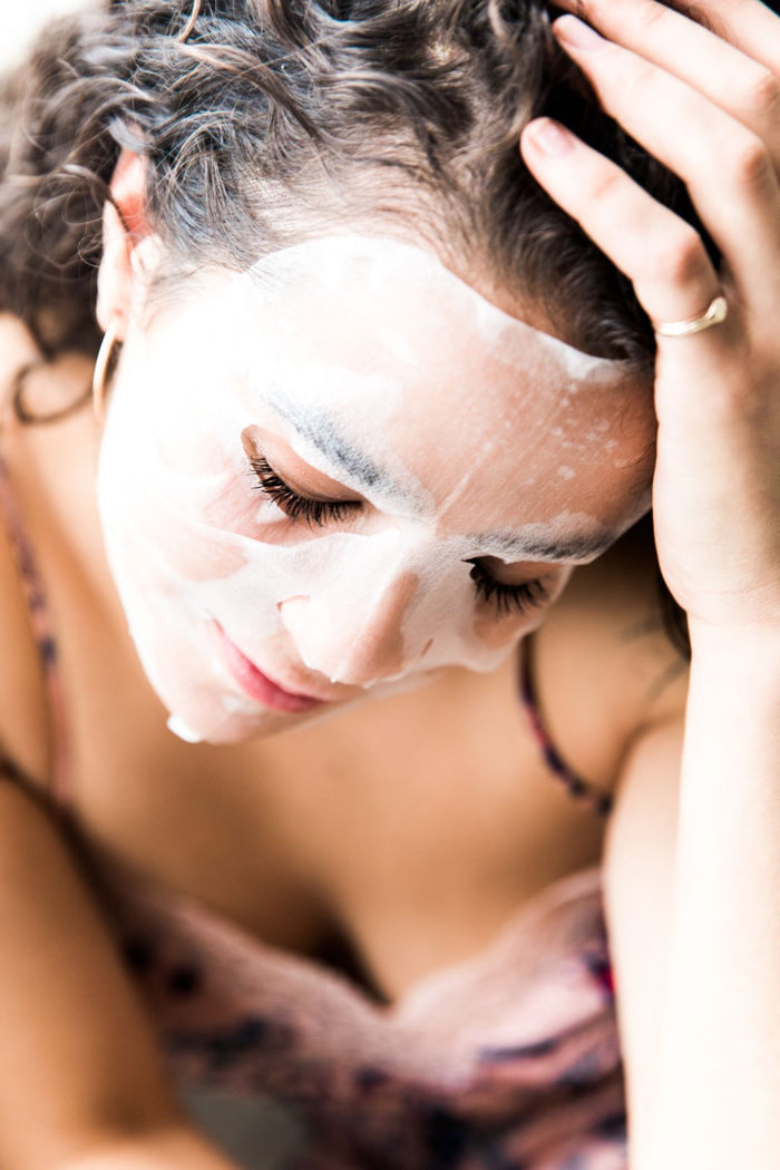 A person with curly hair wears a white ORGAID Anti-Aging & Moisturizing Organic Sheet Mask, sitting serenely with closed eyes and a hand on their head, dressed in a spaghetti strap top, indulging in a skincare ritual.