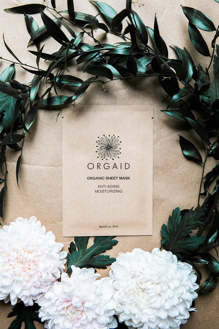 An Anti-Aging & Moisturizing Organic Sheet Mask by ORGAID elegantly rests among dark green leaves and white flowers on a brown surface, offering anti-aging benefits and superior moisturizing care.