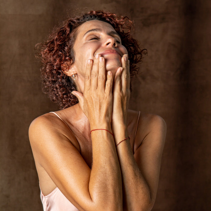A woman with curly hair wears a sleeveless top and smiles gently, touching her face with both hands. She looks relaxed and content against a brown background, possibly enjoying the benefits of Tree To Tubs Anti-Aging Peptide & Niacinamide Serum For Face.