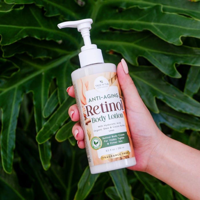 A hand holds a bottle of Tree To Tub Anti Aging Retinol Body Lotion with Shea & Cocoa Butter against lush green leaves. This fragrance-free lotion, enriched with hyaluronic acid and organic shea cocoa butter, addresses fine lines and deep wrinkles.
