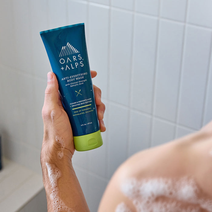 A person with soapy skin holds a tube of Oars + Alps Anti-Everything Body Wash in a shower with white tiled walls.