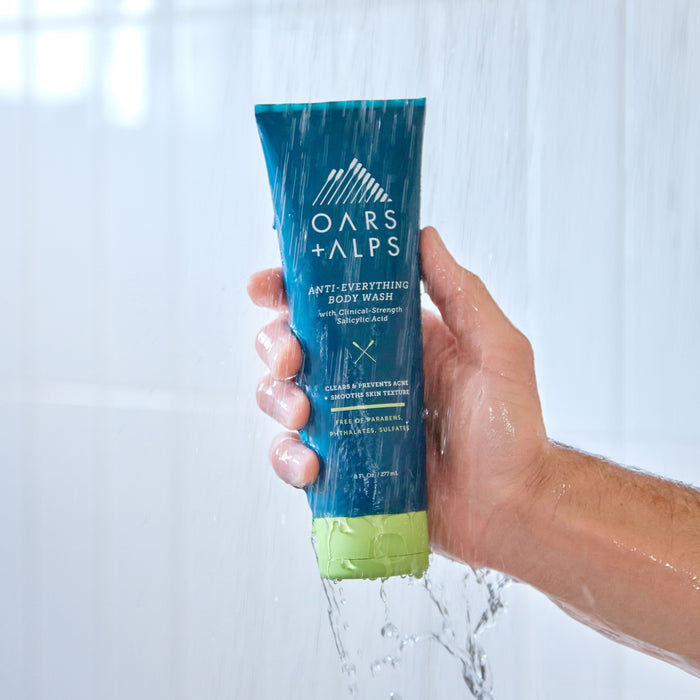 A hand holds a blue and green tube of Oars + Alps Anti-Everything Body Wash under running shower water against a white tiled background.