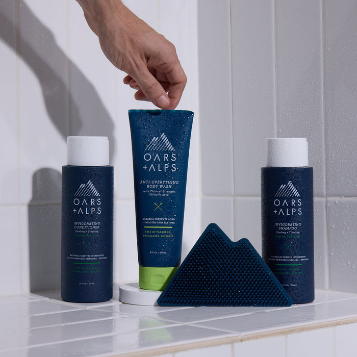 A hand picks up a blue tube of Oars + Alps Anti-Everything Body Wash from a tiled shower ledge, where two bottles of shampoo, conditioner, and a blue textured scrubber are also placed.