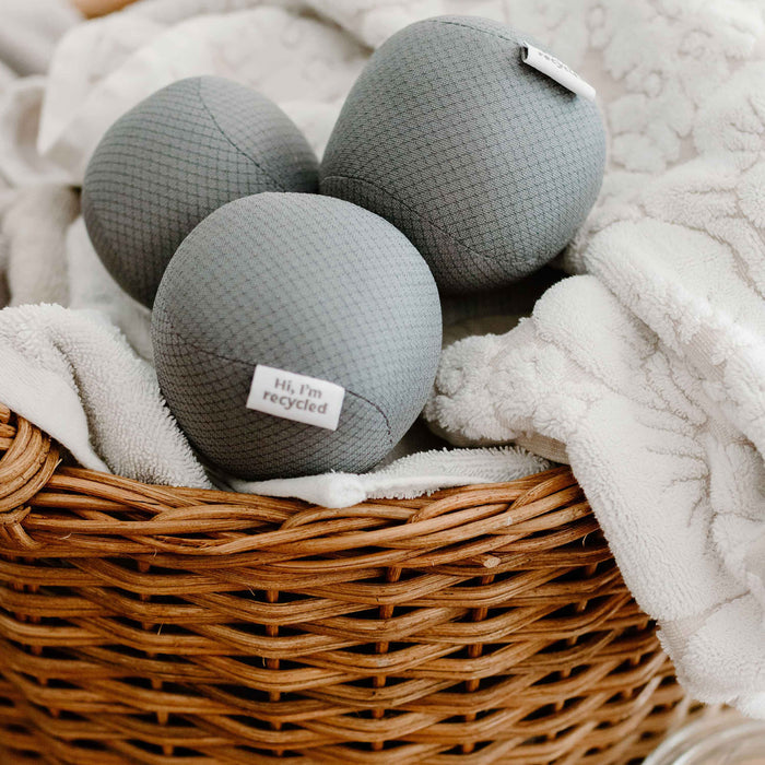 A wicker basket contains a soft white blanket and three gray dryer balls. Each ball from Full Circles Anti-Static Dryer Balls highlights an eco-friendly laundry solution with labels reading, Hi, Im recycled, promising wrinkle-free clothes.