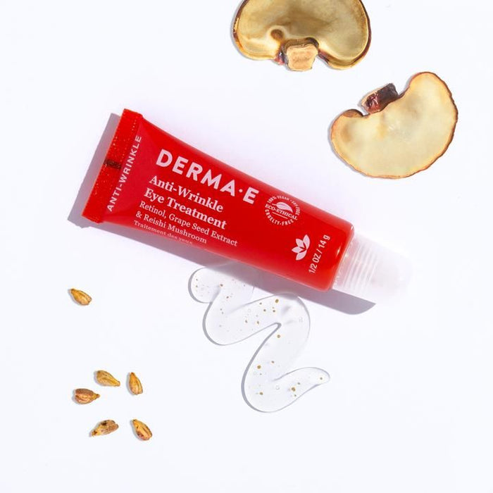 A red tube of Derma E Anti-Wrinkle Eye Treatment Gel sits on a white surface, showcasing its Retinol and Reishi Mushroom Extracts. Mushroom slices, grape seeds, and clear gel are artfully arranged around it.