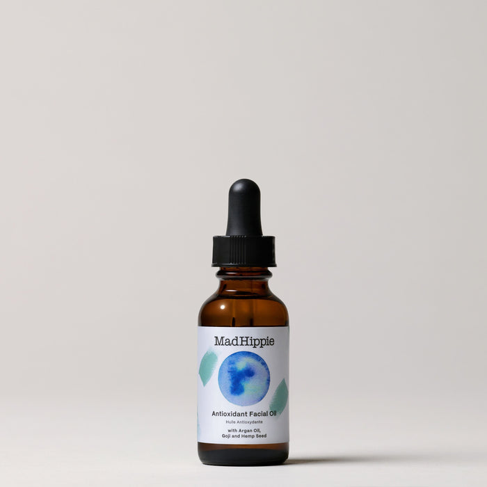 A Mad Hippie Antioxidant Facial Oil brown glass dropper bottle is set against a light gray background, highlighting its natural ingredients for skin hydration. The label displays a blue and green circular design.