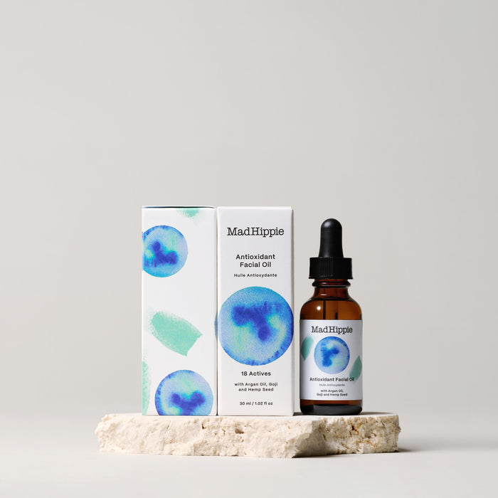A brown dropper bottle labeled Antioxidant Facial Oil from Mad Hippie stands on a stone slab next to its matching package. Featuring abstract blue and green watercolor designs, the box showcases the oils skin hydration benefits and natural ingredients on a light background.