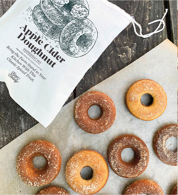 A FarmSteady Apple Cider Doughnut Baking Mix bag sits on a wooden surface, offering a taste of homemade treats. Below, doughnuts made from the mix, some with cinnamon-sugar coating and others plain, are arranged neatly on parchment paper.