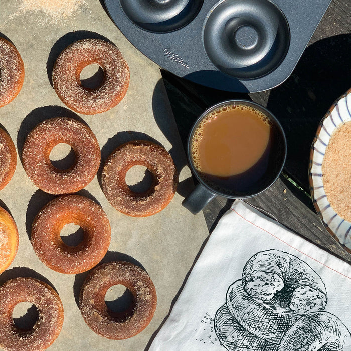 FarmSteadys Apple Cider Doughnut Baking Mix creates freshly baked doughnuts, perfectly dusted with cinnamon-sugar. These are presented on a baking sheet next to a steaming coffee mug and a doughnut pan, with a charming Apple Cider Doughnut tea towel accentuating the wooden surface.