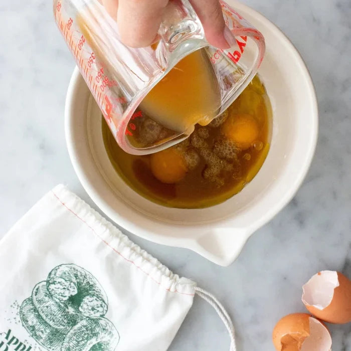 A hand pours apple cider from a measuring cup into a bowl with eggs and sugar, the first step in preparing Apple Cider Doughnuts with FarmSteady Apple Cider Doughnut Baking Mix. Eggshells and a cloth bag rest nearby on a marble surface.
