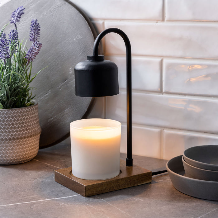 The Arched Candle Warmer Lamp by Candle Warmers Etc. features a modern design with top-down warming technology. It displays a white candle on a sleek wood base, accompanied by a plant in a patterned pot and gray bowls, all against an elegant white tiled wall.