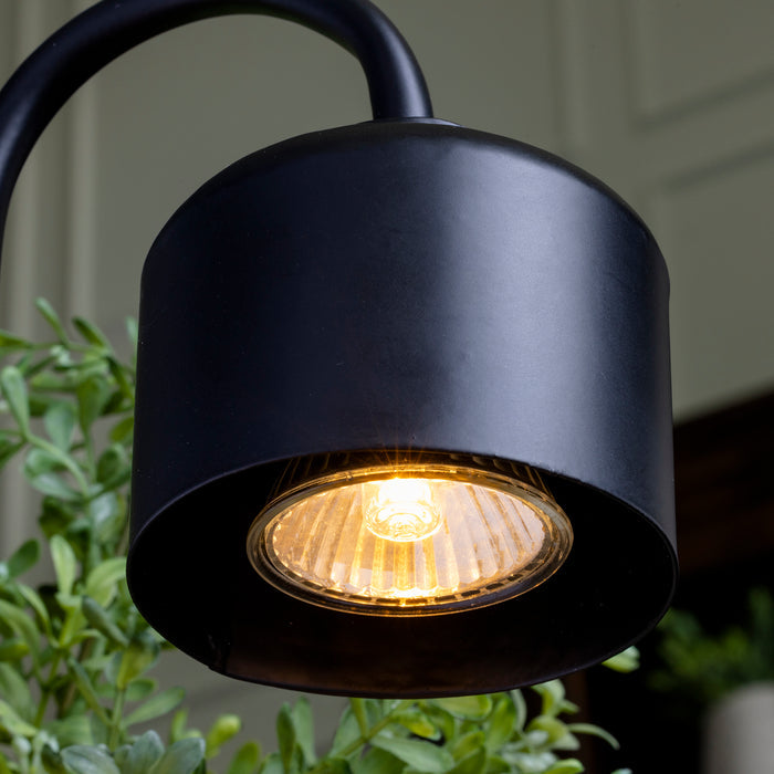 The Arched Candle Warmer Lamp by Candle Warmers Etc., featuring a black metal design and wooden base, casts a warm glow. Surrounded by greenery, it enhances any garden or patio with its inviting blend of nature and design.