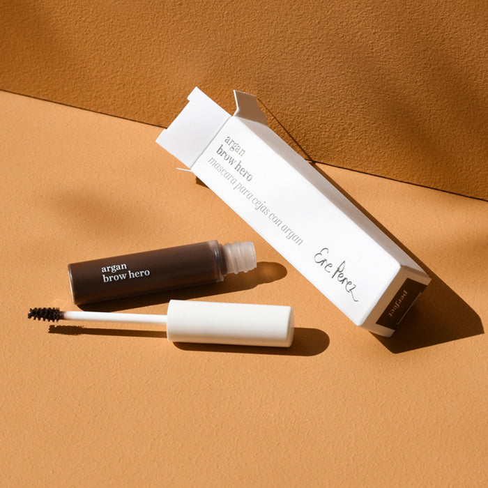 On a light brown surface, Ere Perezs Argan Brow Hero brow gel tube with vegan fibers is displayed beside its open white box, accompanied by a nearby brush wand, all softly shadowed to enhance the scene.