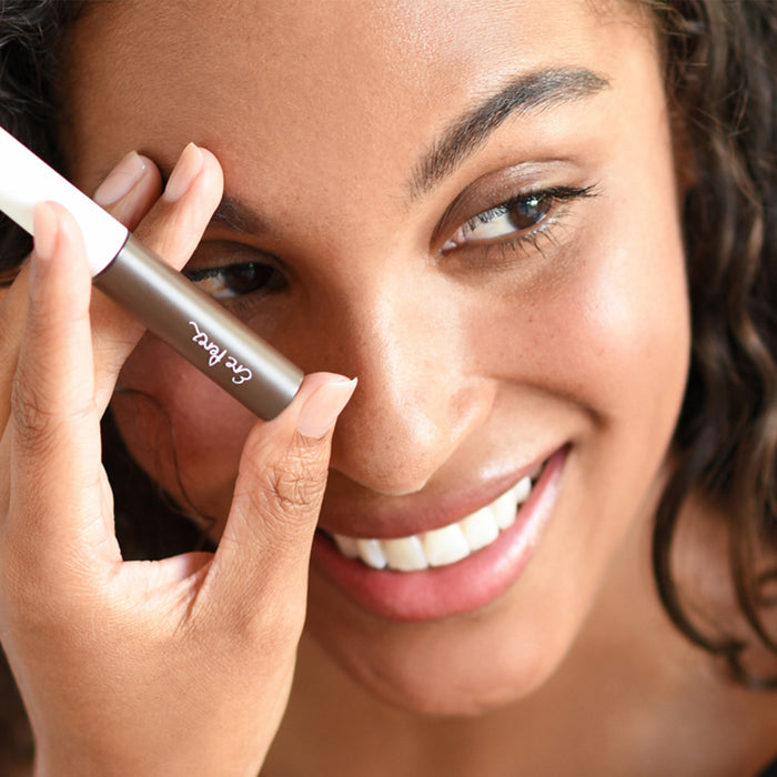A woman smiles while expertly shaping her brows with Ere Perezs Argan Brow Hero. Her clear skin and curly hair frame her face beautifully. The brush, with its white and brown handle, holds tinted gel infused with vegan fibers, adding the perfect touch to her look.