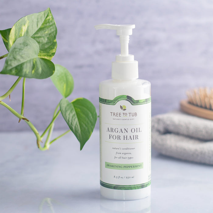 A bottle of Tree To Tubs Argan Oil Cleansing Conditioner for Oily Hair with a pump rests on a surface beside folded gray towels and a wooden hairbrush, with a green leafy plant against a marble-like wall in the background.