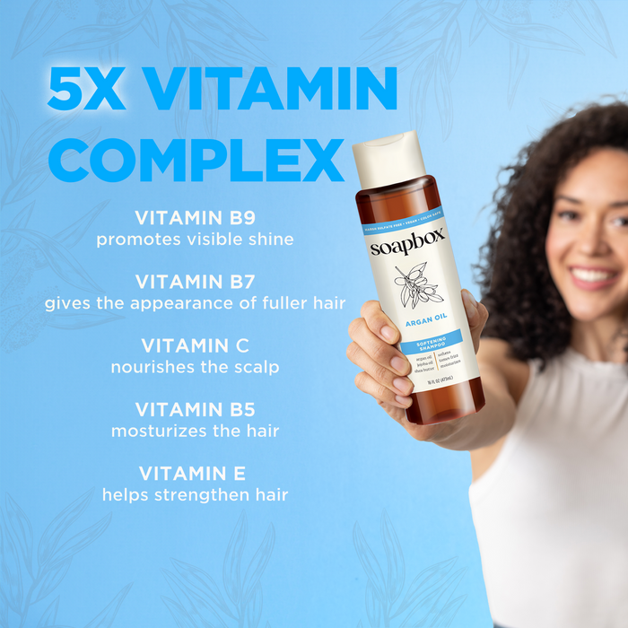 A woman with curly hair holds Soapboxs Argan Oil Softening Shampoo against a blue backdrop. The text highlights its vitamin complex (B9, B7, C, B5, E), promoting shine, fullness, nourishment, moisture, and strength for healthy hair.