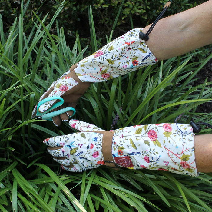 A person wearing Womanswork Arm Saver Gloves with leather palms and floral patterns uses teal scissors to trim green plants, surrounded by lush greenery, showcasing the elegance and functionality of these quality gardening gloves.