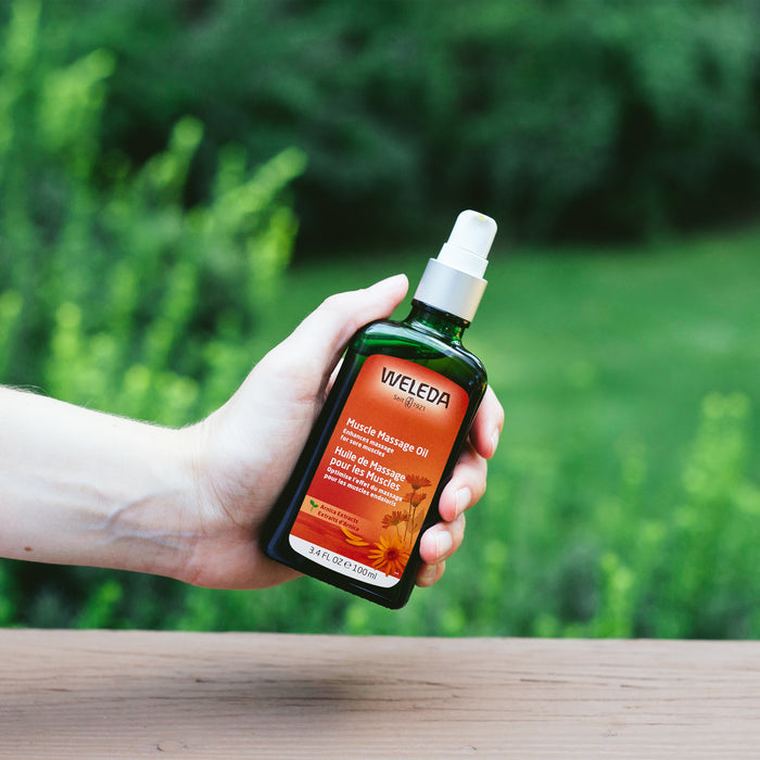A hand holds Weledas Arnica Muscle Massage Oil outdoors, where the orange and yellow label with pump contrasts against the green foliage, highlighting its natural arnica flower-derived essential oils.