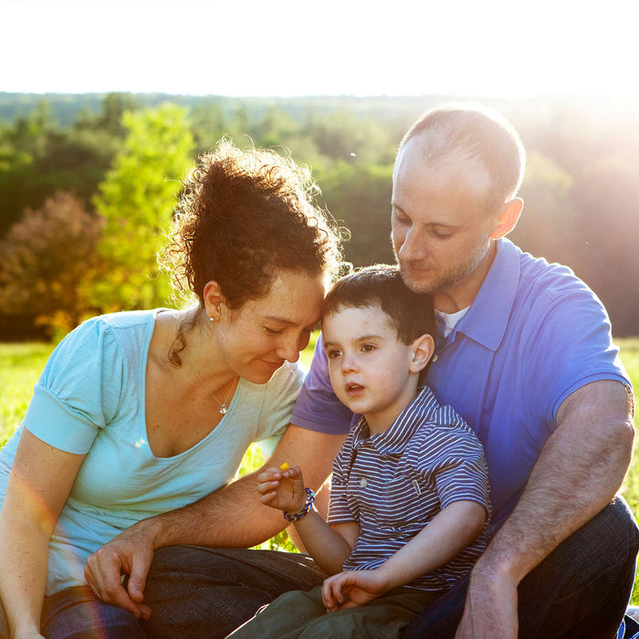 A family enjoys a sunny day outdoors, surrounded by the scent of Badger Balms Aromatic Chest Rub. A curly-haired woman and a bearded man lovingly embrace a young boy between them, with trees and sunlight adding warmth to the tranquil scene.