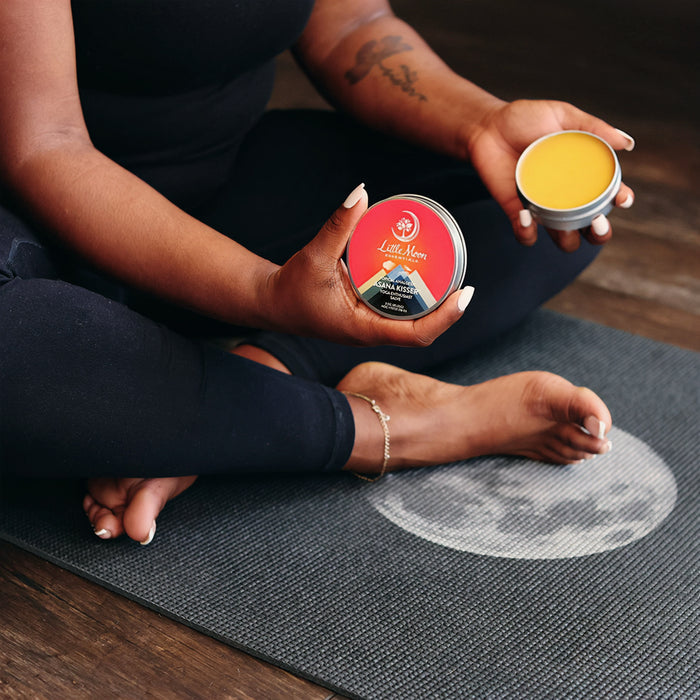 A yoga enthusiast sits cross-legged on a mat with an open container of soothing yellow Asana Kisser™ Pain Relief Salve by Little Moon Essentials in hand. Their tattooed arm, black clothing, and ankle bracelet complete the scene, alongside another container with a red label.