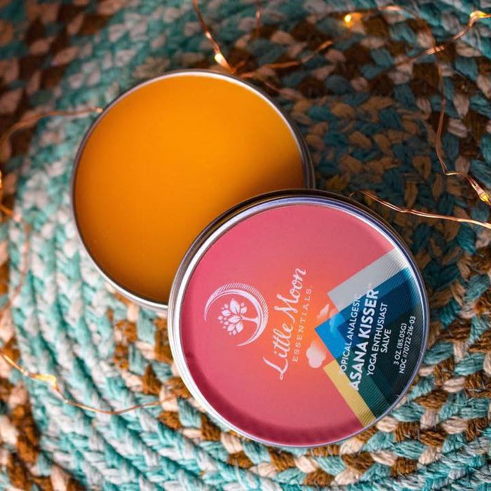 An open tin labeled Little Moon Essentials, Asana Kisser™ - Pain Relief Salve, is on a woven mat with orange balm exposed—ideal for yoga lovers. Small twinkle lights encircle the background, creating a calming ambiance for soothing sore muscles and relieving pain.