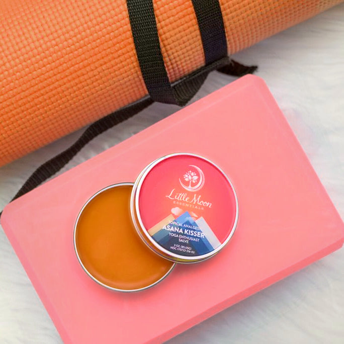 An open tin of Asana Kisser™ Pain Relief Salve by Little Moon Essentials reveals its orange, natural balm on a pink yoga block. In the background, an orange yoga mat with a black strap awaits dedicated yoga enthusiasts.