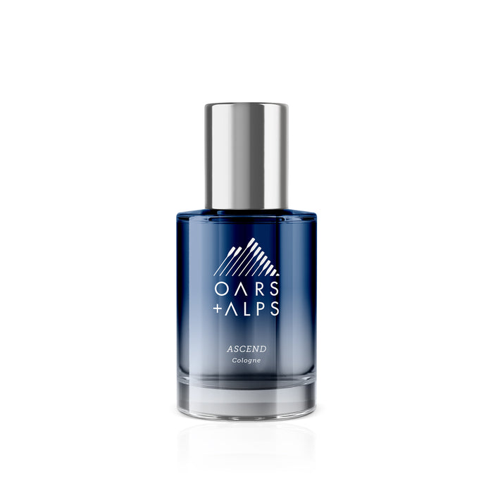A blue glass bottle of Oars + Alps ASCEND Cologne with a silver cap features a white mountain logo and text on the front, set against a white background—a captivating cedar-scented fragrance by Oars + Alps.