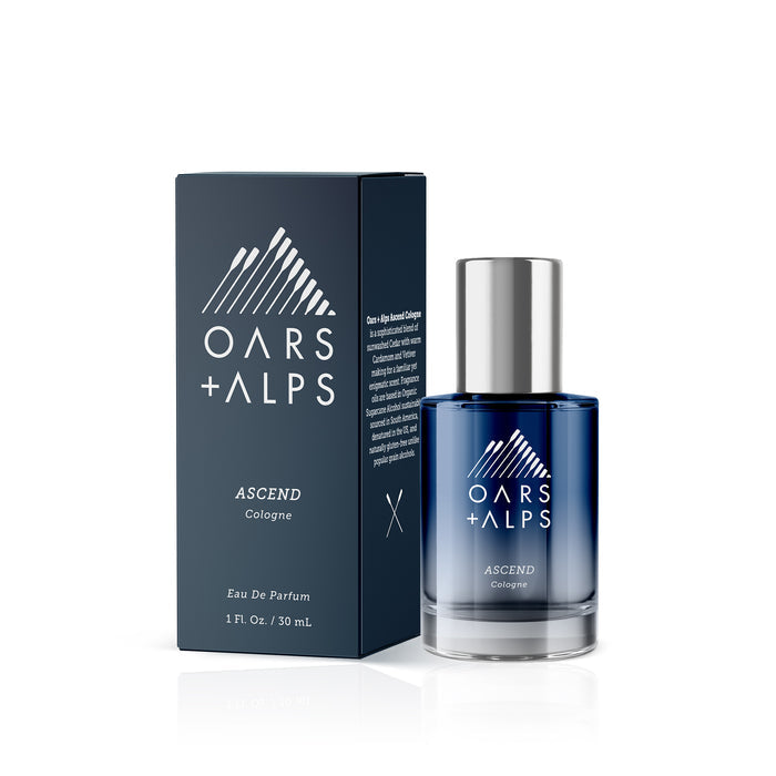A 30 mL dark blue and silver bottle of Oars + Alps ASCEND Cologne with an alpine-inspired scent stands beside its matching blue box featuring white mountain graphics.