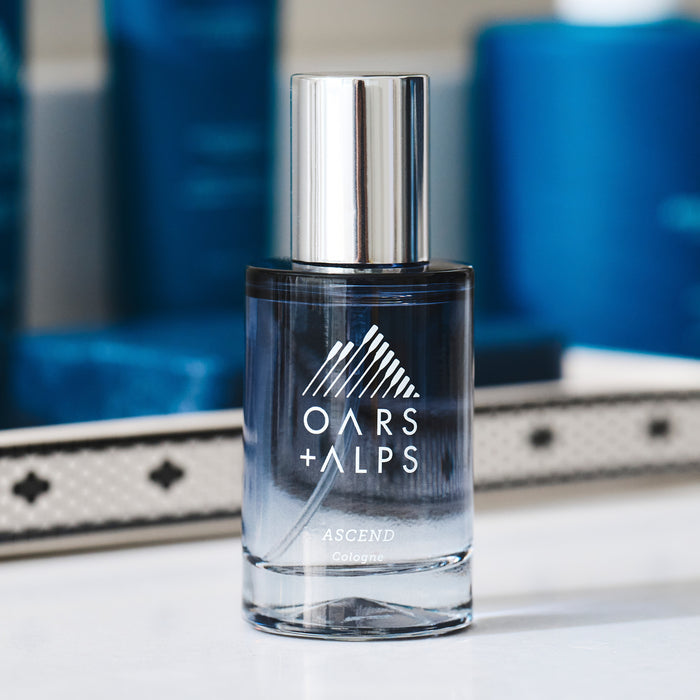 A bottle of Oars + Alps ASCEND Cologne with a silver cap rests on a white surface, its allure suggested by a blurred blue background and the glint of a nearby decorative mirror.