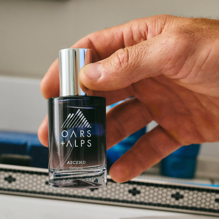 A hand holds a bottle of Oars + Alps ASCEND Cologne with a silver cap, its enticing scent ready to impress, set against a bathroom countertop.