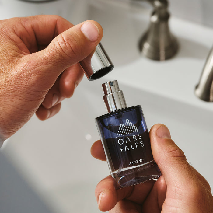 A person holds a bottle of Oars + Alps ASCEND Cologne and removes the silver cap, getting ready to use it by the bathroom sink.