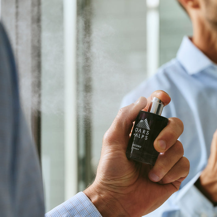 A person in a blue shirt sprays a bottle of Oars + Alps ASCEND Cologne in front of a mirror, the mist of fragrance and its sophisticated blend lingering in the air.