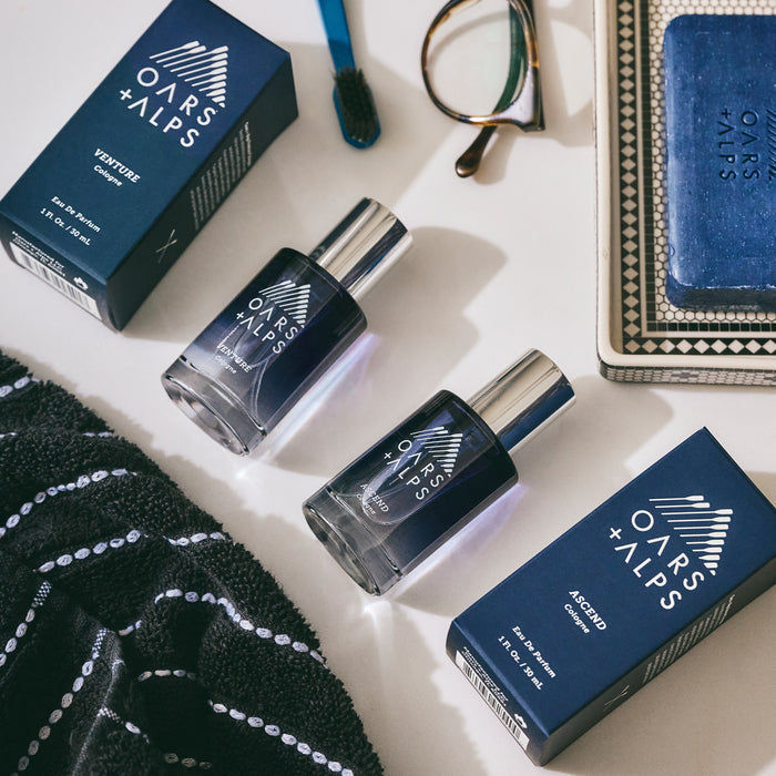 Oars + Alps ASCEND Cologne in its blue bottle and box sits on a bathroom counter beside a toothbrush, eyeglasses, blue soap, and a black-and-white striped towel—poised to deliver an enigmatic scent with every spray.