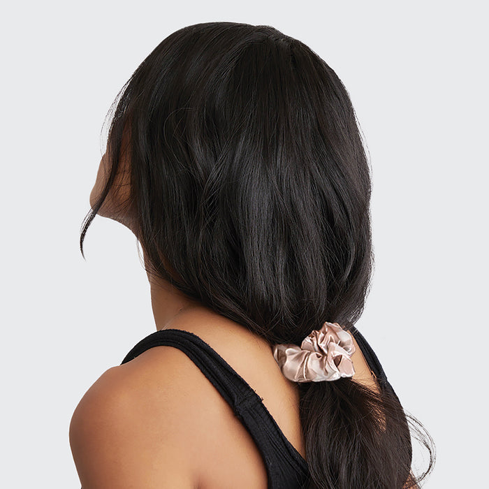 An individual with long, dark hair faces away from the camera, their locks secured in a ponytail with a light pink Kitsch Assorted Satin Sleep Scrunchie. This chic accessory keeps their hair frizz-free and prevents breakage. They wear a black sleeveless top against a plain background.