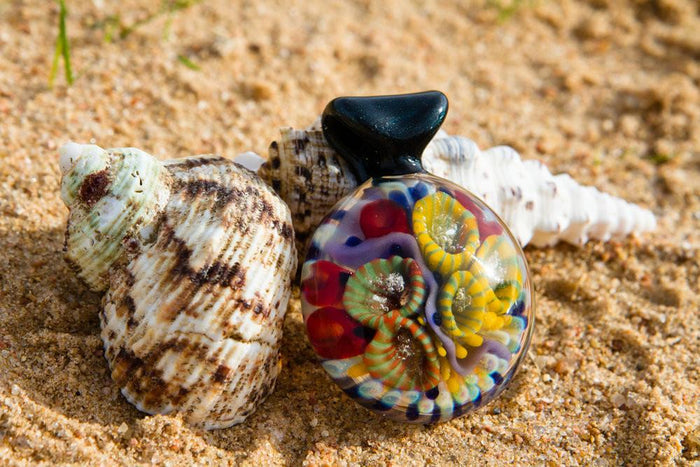 Round glass pendant with a colorful coral reef interior containing cremains, set on sand.