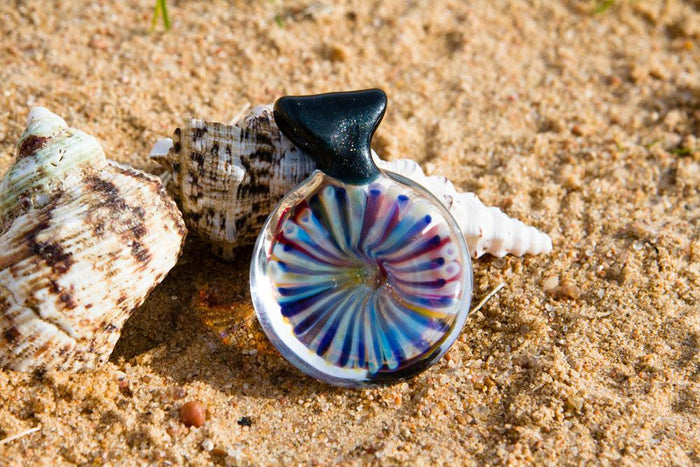 Round glass pendant showing a vivid multicolored coral reef interior with cremains, photographed on sand.