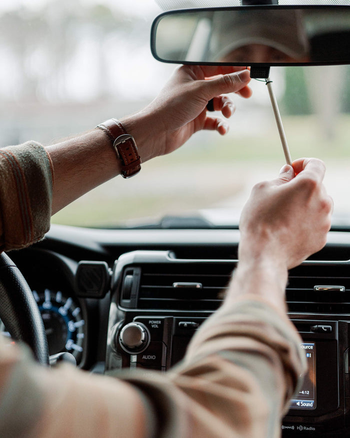 Sitting in a car with audio and climate controls, a person adjusts the rearview mirror, their brown watch complementing a plaid shirt. They enjoy the fresh scent from Enviroscents Auto Air Freshener Stix as trees and road unfold through the windshield.